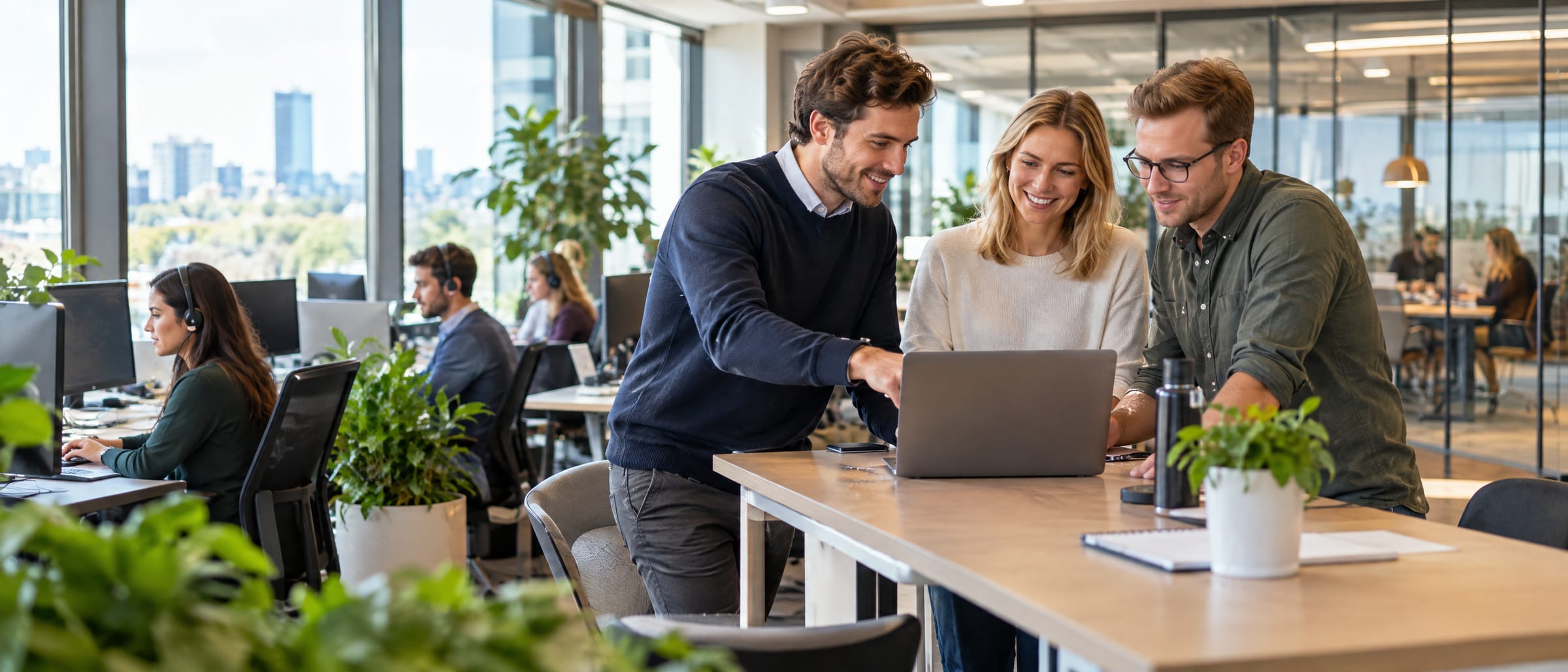 Three colleagues talking over a laptop in a bright open-plan office, with teammates at their desks behind